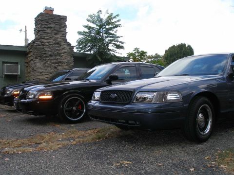 1998 Ford Crown Victoria - Two dark blue/black Ford Crown Victoria sedans parked at an angle on a paved driveway, with the foreground car showing a Ford grille badge,…