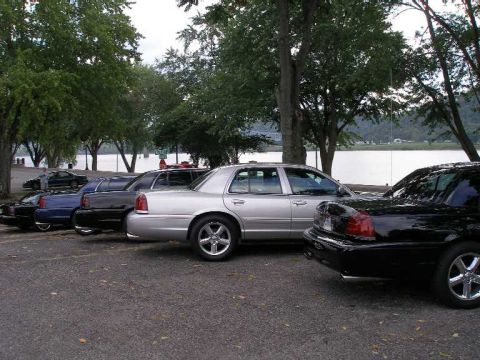 1998 Ford Crown Victoria - A row of Ford/Mercury full-size sedans lined up at a waterfront park, a silver Crown Victoria on chrome wheels in the center flanked by dar…