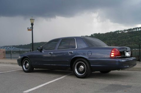 1998 Ford Crown Victoria - A dark blue Ford Crown Victoria sedan parked side-on in a riverfront lot under heavy storm clouds, wearing black steel wheels with chrome c…