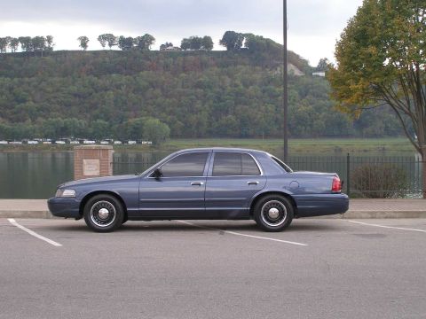 1998 Ford Crown Victoria - Slate blue Ford Crown Victoria sedan in full driver-side profile in an empty riverfront parking lot, wearing black steel wheels with police…
