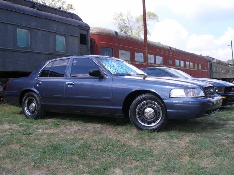 1998 Ford Crown Victoria - A blue Ford Crown Victoria sedan parked on grass in front of vintage railroad passenger cars, one lettered PENNSYLVANIA. It wears black ste…
