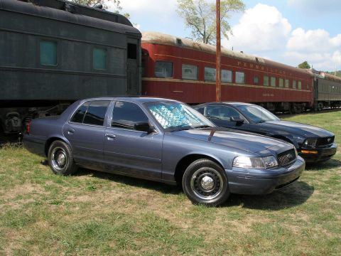 1998 Ford Crown Victoria - A blue-gray Ford Crown Victoria sedan with steel wheels and hood stripes parked on grass in front of vintage railroad passenger cars, one l…