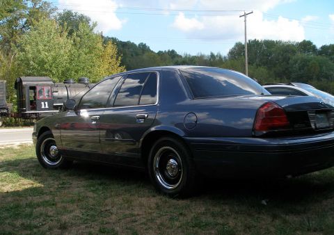 1998 Ford Crown Victoria - Rear three-quarter view of a dark blue Crown Victoria sedan on grass, showing red tail lamps, a fuel door on the left quarter and black ste…