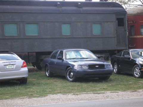 1998 Ford Crown Victoria - A dark blue-gray Ford Crown Victoria sedan parked on grass in front of a black vintage railroad passenger car at a train museum, seen from…