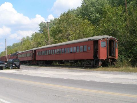 1998 Ford Crown Victoria - A string of derelict maroon passenger railcars parked on a siding beside a rural two-lane road, with a black Jeep Wrangler and a flatbed tr…