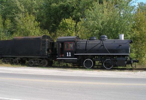 1998 Ford Crown Victoria - A black steam locomotive numbered 11 with white-rimmed driving wheels and a red-framed cab window sits coupled to its tender on a siding be…