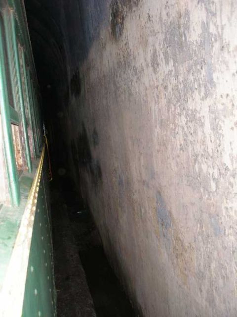 1998 Ford Crown Victoria - Dim view down a narrow gap between a green riveted steel girder structure and a stained concrete tunnel wall, receding into darkness, a pas…