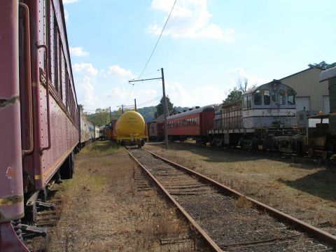 1998 Ford Crown Victoria - Railroad museum yard on a sunny day, looking down weed-grown track between rows of stored rolling stock: a maroon wooden passenger car in t…