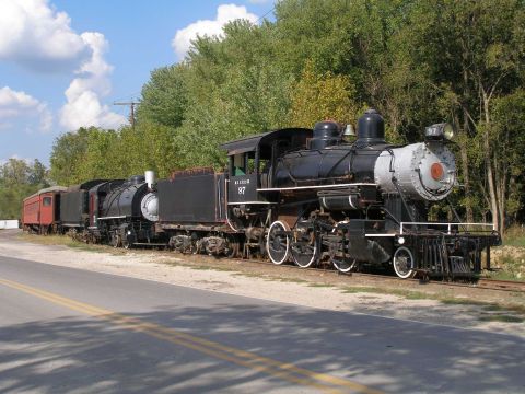 1998 Ford Crown Victoria - A black steam locomotive numbered 97 sits on a siding beside a paved road, coupled to a tender, a second steam engine, and a maroon wooden…