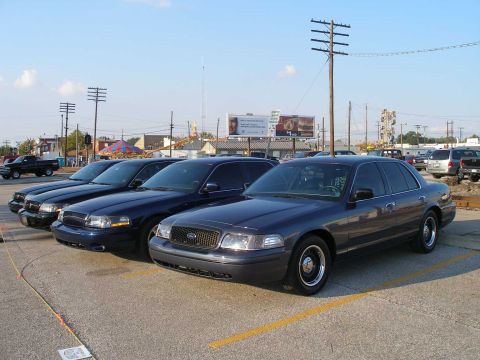 1998 Ford Crown Victoria - A row of three dark blue Ford and Mercury Panther-platform sedans lined up nose-out in a parking lot at an outdoor event, led by a Crown Vi…