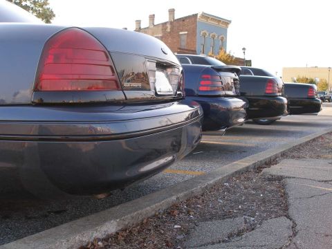 1998 Ford Crown Victoria - Low rear-corner view down a row of dark full-size Ford Panther-platform sedans lined up nose-to-tail in angled street parking, the nearest…