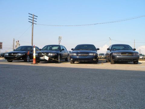 1998 Ford Crown Victoria - Four dark Ford Panther-platform sedans parked nose-out in a row at an outdoor meet, including Mercury Marauder-style cars with waterfall gr…