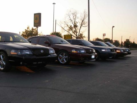 1998 Ford Crown Victoria - A row of full-size Mercury and Ford sedans parked diagonally in a lot at sunset, led by a black car with chrome five-spoke wheels and a Mer…