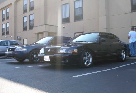1998 Ford Crown Victoria - Two dark Mercury sedans parked nose-out in front of a tan hotel building; the foreground car is a black full-size sedan lowered on polished…