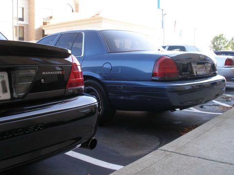 1998 Ford Crown Victoria - Rear three-quarter view of a dark blue Ford Crown Victoria sedan parked in a lot, with a black Mercury Marauder in the foreground showing i…