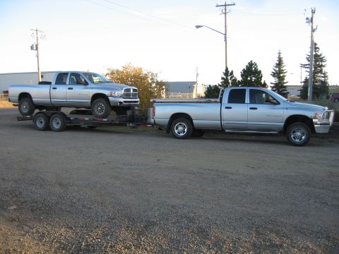 2005 Dodge 3500 - A silver 2005 Dodge 3500 pickup truck tows a second identical truck on a trailer under a clear sky.