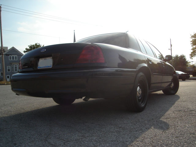 Rear quarter view of a black sedan parked on asphalt. Another car is visible in the distance