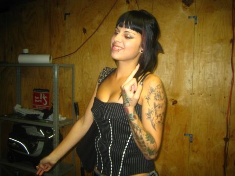 1968 Ford Mustang - Woman with extensive tattoos poses in a corset, with shelves and a security sign visible in the background.