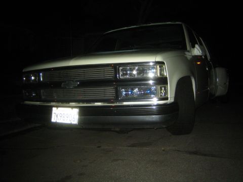1990 Ford Bronco - Low-angle night view of the front corner of a white Chevrolet dually pickup with a bowtie emblem and dual rear wheels.