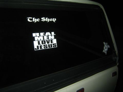 1990 Ford Bronco - vehicle's rear window at night displays white decals reading The Shop and REAL MEN LOVE JESUS beside a hibiscus-turtle sticker.