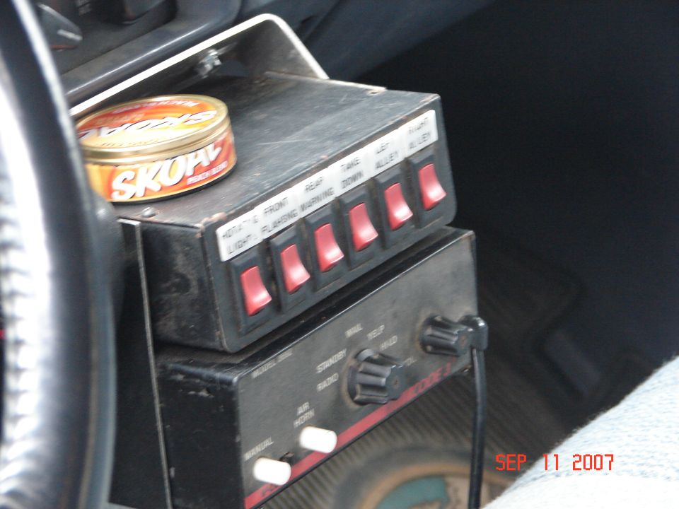 Six-switch emergency light control box and Code 3 siren unit inside a vehicle, with a Skoal tin on top.