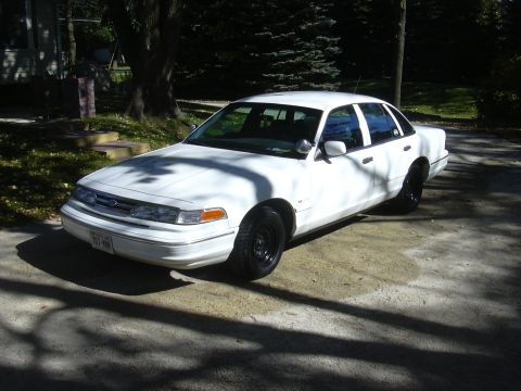 1997 Ford Crown Victoria - A white sedan with black wheels is parked on a paved surface, with trees in the background.