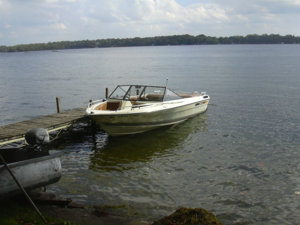 White runabout powerboat moored to wooden dock on a calm lake with wooded shoreline.