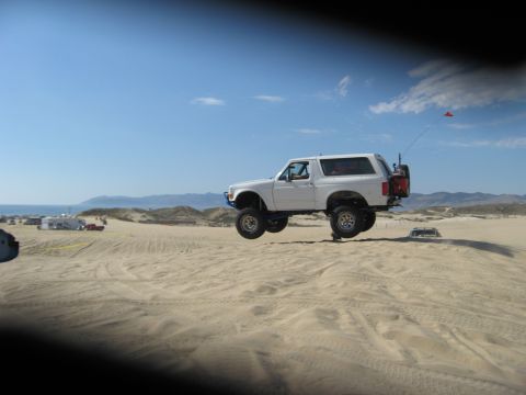 1994 Ford Bronco - A white 1994 Ford Bronco soars through the air over sand dunes on a clear day.