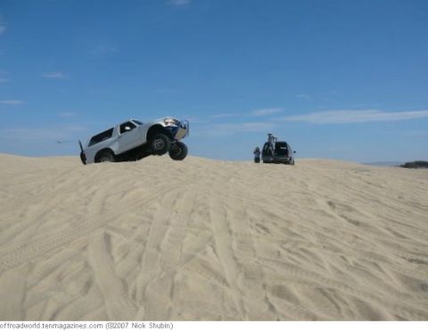 1994 Ford Bronco - Two vehicles navigate sandy terrain under a clear blue sky, with one SUV cresting a dune and another stopped with people on its roof.