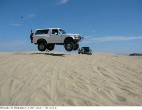 1994 Ford Bronco - A white Ford Bronco jumps over sand dunes with another vehicle in the background under a clear blue sky.