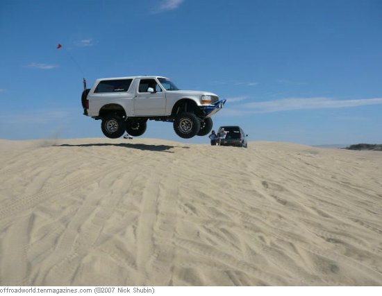 White Ford Bronco airborne over sand dunes, with a dark SUV behind it