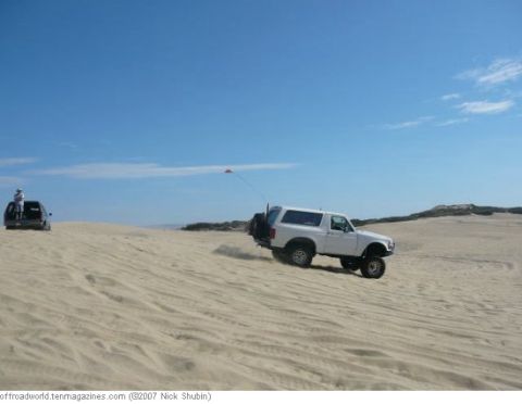 1994 Ford Bronco - A white 1994 Ford Bronco navigates sandy terrain under a clear blue sky, with another vehicle visible in the distance.
