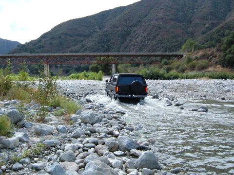 1981 Ford Bronco - A black Ford Bronco navigates a shallow river with a bridge overhead.