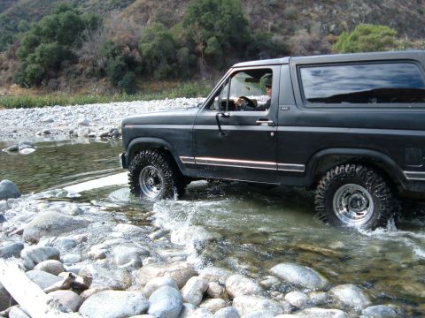 1981 Ford Bronco - A dark 1981 Ford Bronco navigates a rocky stream bed, kicking up water.