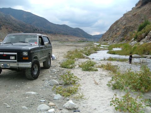 1981 Ford Bronco - A person stands in a shallow river in a rocky, mountainous canyon next to a parked SUV.