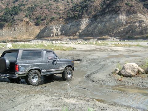 1981 Ford Bronco - A dark gray Ford Bronco navigates a muddy off-road course with rocky hills in the background.