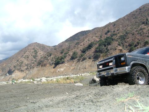1981 Ford Bronco - This black SUV navigates a rocky, off-road trail with steep, arid mountains looming behind it.