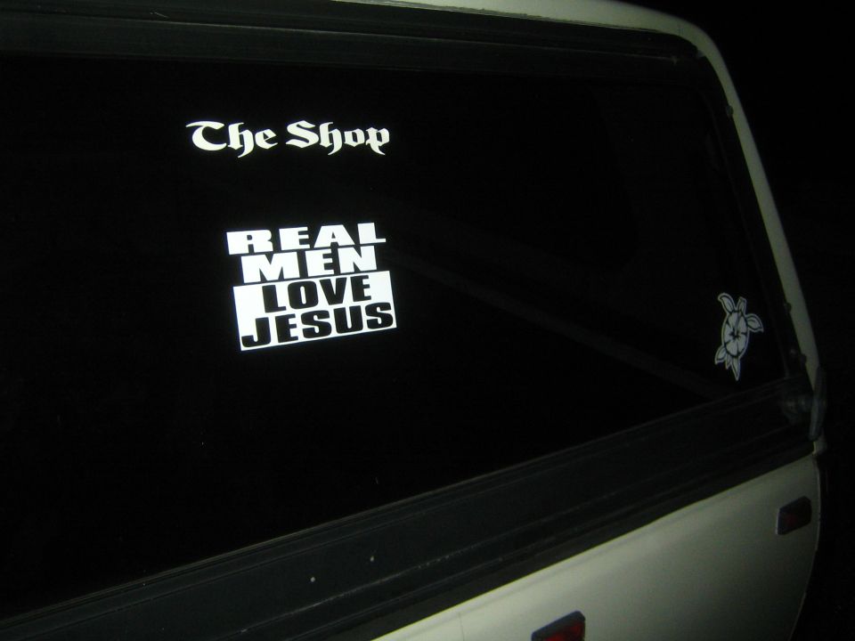 White truck's rear window with 'The Shop' and 'REAL MEN LOVE JESUS' stickers
