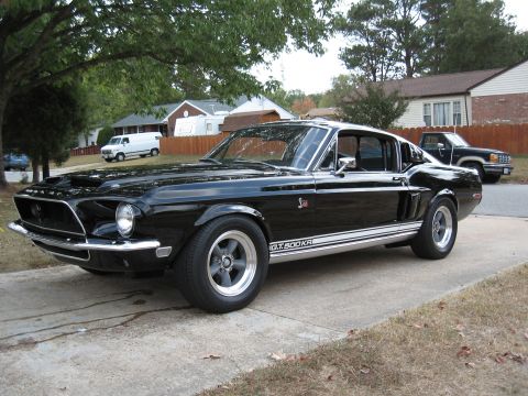 1968 Shelby GT500 - A black 1968 Shelby GT500KR coupe is parked on a driveway with houses and trees in the background.