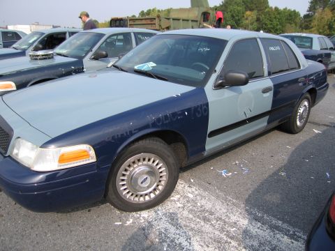 1991 Ford LTD Crown Victoria - A two-tone police car is parked in a lot with other vehicles and debris on the ground.