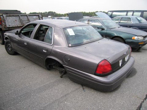 1991 Ford LTD Crown Victoria - A dark gray sedan, missing its rear wheels, sits in a lot with other vehicles and a dump truck.