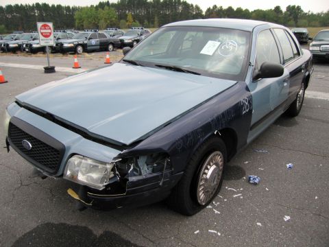 1991 Ford LTD Crown Victoria - Damaged police car with a flat tire and visible damage to the front bumper and headlight. Multiple similar vehicles are parked in the backg…