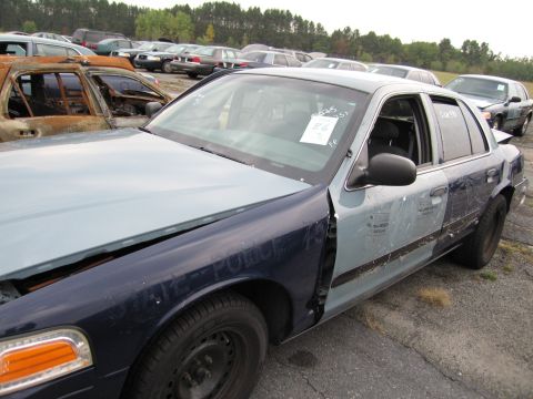 1991 Ford LTD Crown Victoria - This damaged police car sits in a junkyard, surrounded by other vehicles under an overcast sky.