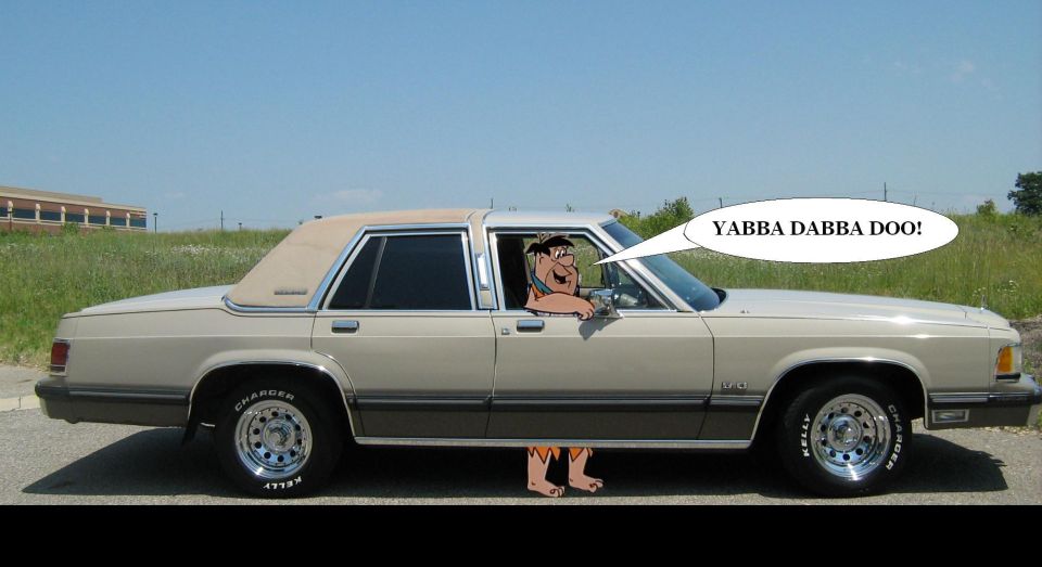Side view of a beige 1988 Mercury Grand Marquis with Fred Flintstone driving and saying Yabba Dabba Doo!