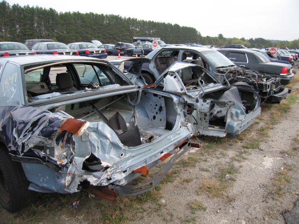 Several wrecked police cars lined up in a junkyard. The closest car is missing its doors and has severe rear-end damage
