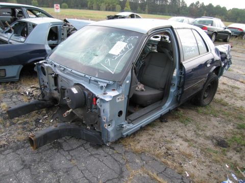 1991 Ford LTD Crown Victoria - A heavily damaged blue sedan sits in a junkyard with its front end missing and windshield cracked.