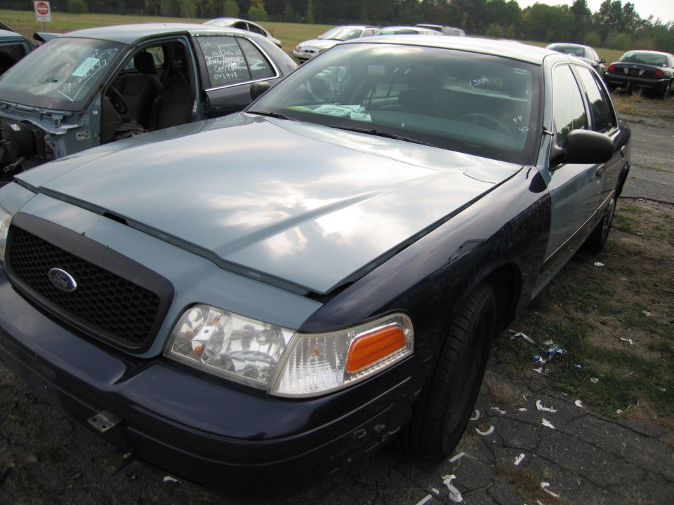 Ford Crown Victoria with two-tone paint, hood open, parked in a junkyard. Another car is visible behind it