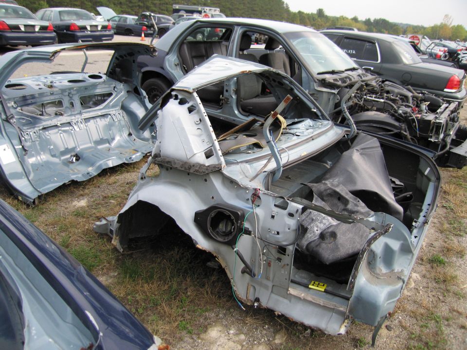 Wrecked light blue car chassis with doors removed, showing interior and engine bay. Multiple other cars in background