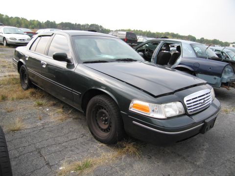1991 Ford LTD Crown Victoria - Dark green sedan in a junkyard, hood ajar, showing a front quarter view. The body style suggests a model from the early 2000s.
