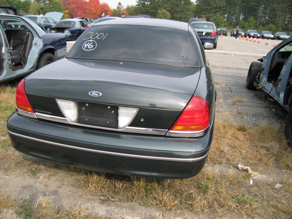 Dark green Ford Crown Victoria, rear view. Numbers 3001 and 46 written on rear window. Another car is partially visible on the left, and a stripped car on the right